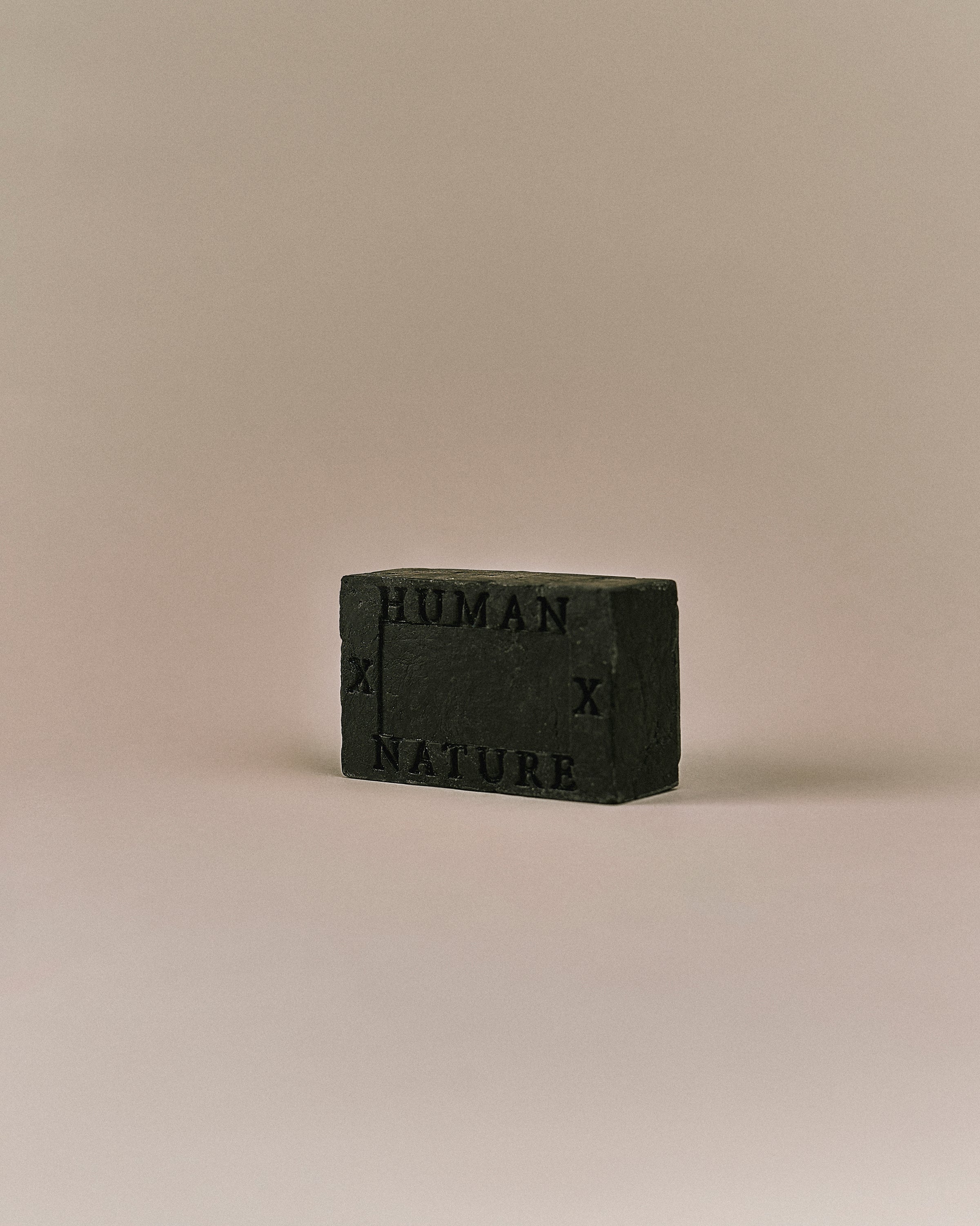 Nhuka Charcoal One Bar shown at an angle with Human x Nature emboss. Handmade in Australia, pH-balanced, sulfate-free, silicone-free, and organic. Warming chai scent with cinnamon, ginger, and nutmeg. Contains pumice for exfoliation. Suitable for hair, face, and body.