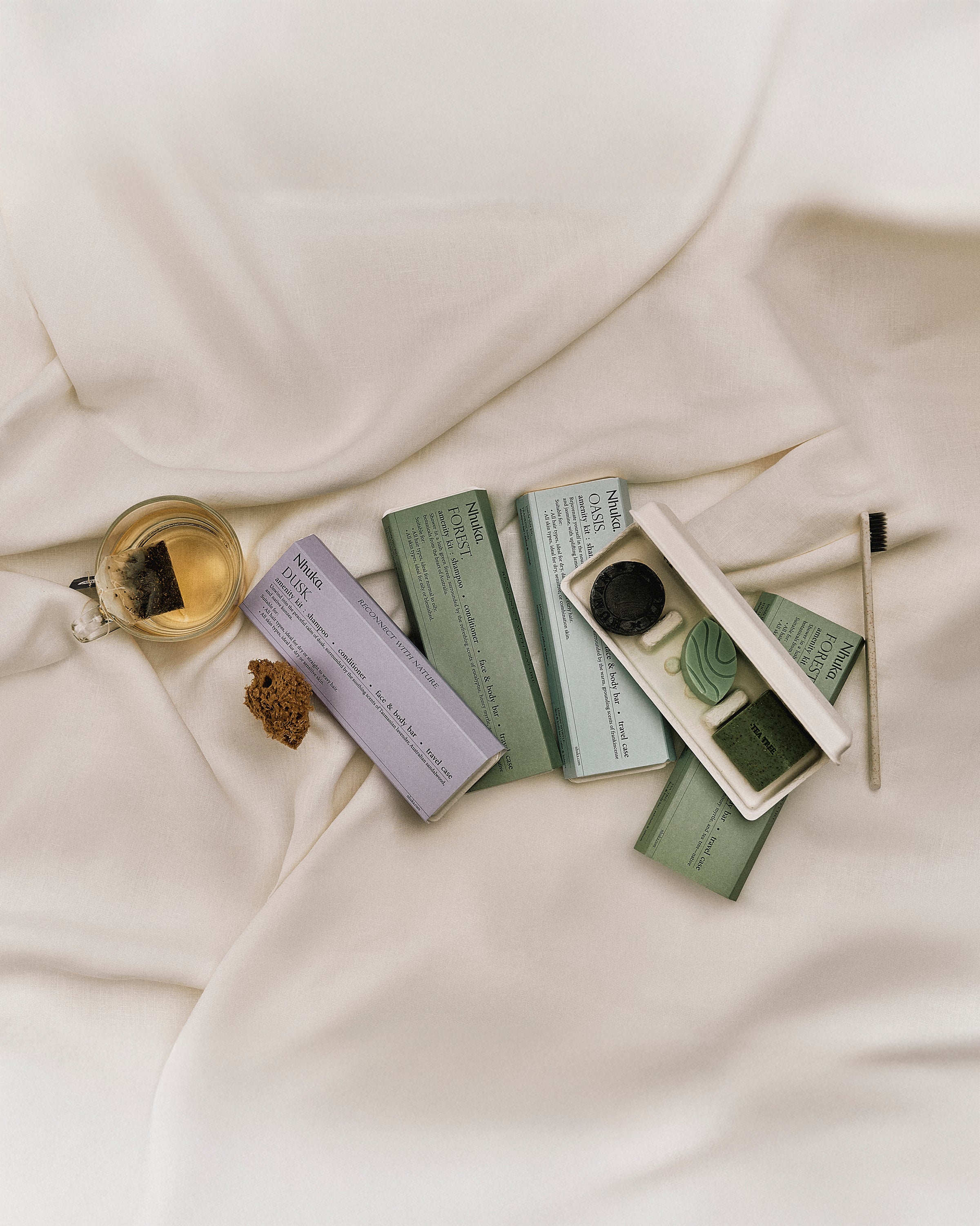Nhuka Amenity Kits displayed on soft white bedding with herbal tea and natural sponge. A calming flat lay capturing the joy of travel and reconnection. Features our Forest, Dusk, and Oasis kits designed for mindful journeys.