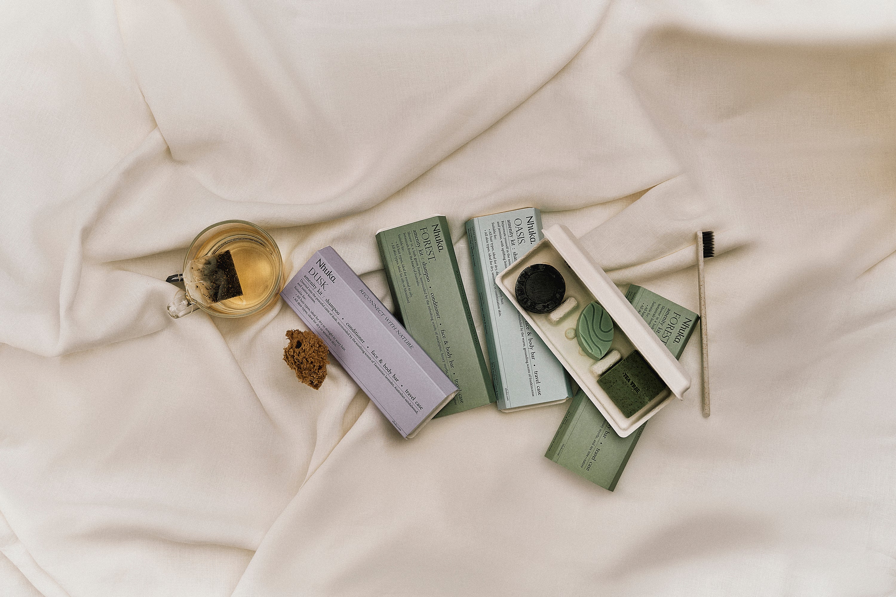 Nhuka Amenity Kits displayed on soft white bedding with herbal tea and natural sponge. A calming flat lay capturing the joy of travel and reconnection. Features our Forest, Dusk, and Oasis kits designed for mindful journeys.