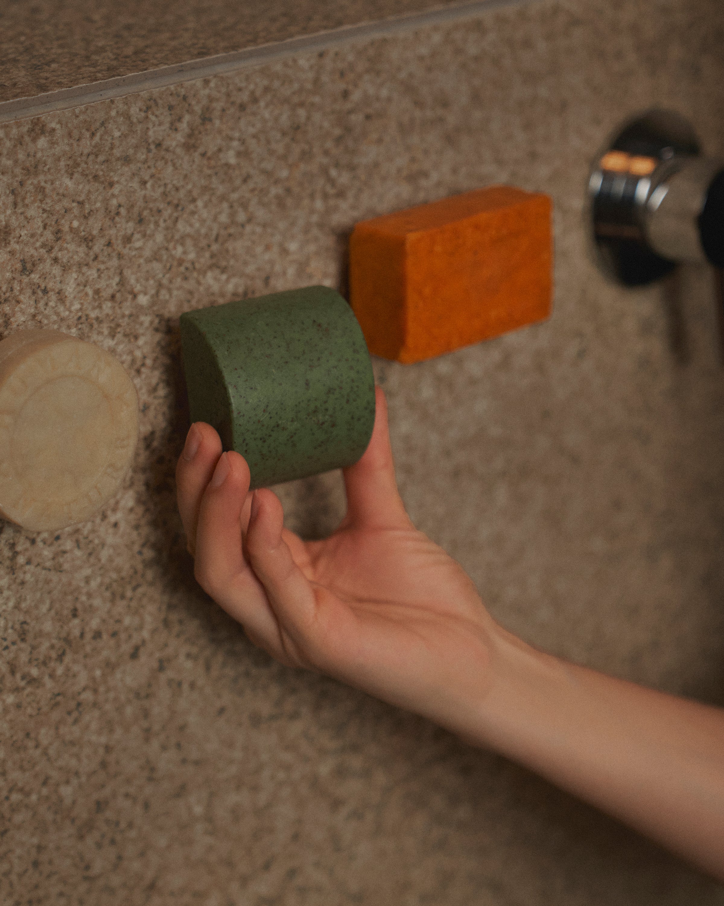 Nhuka Stem magnetic bar holder with shampoo bar, soap bar, and One Bar suspended neatly in the shower. A hand reaches for the bar, evoking restfulness and simplicity. Highlights our Shower Essentials collection.