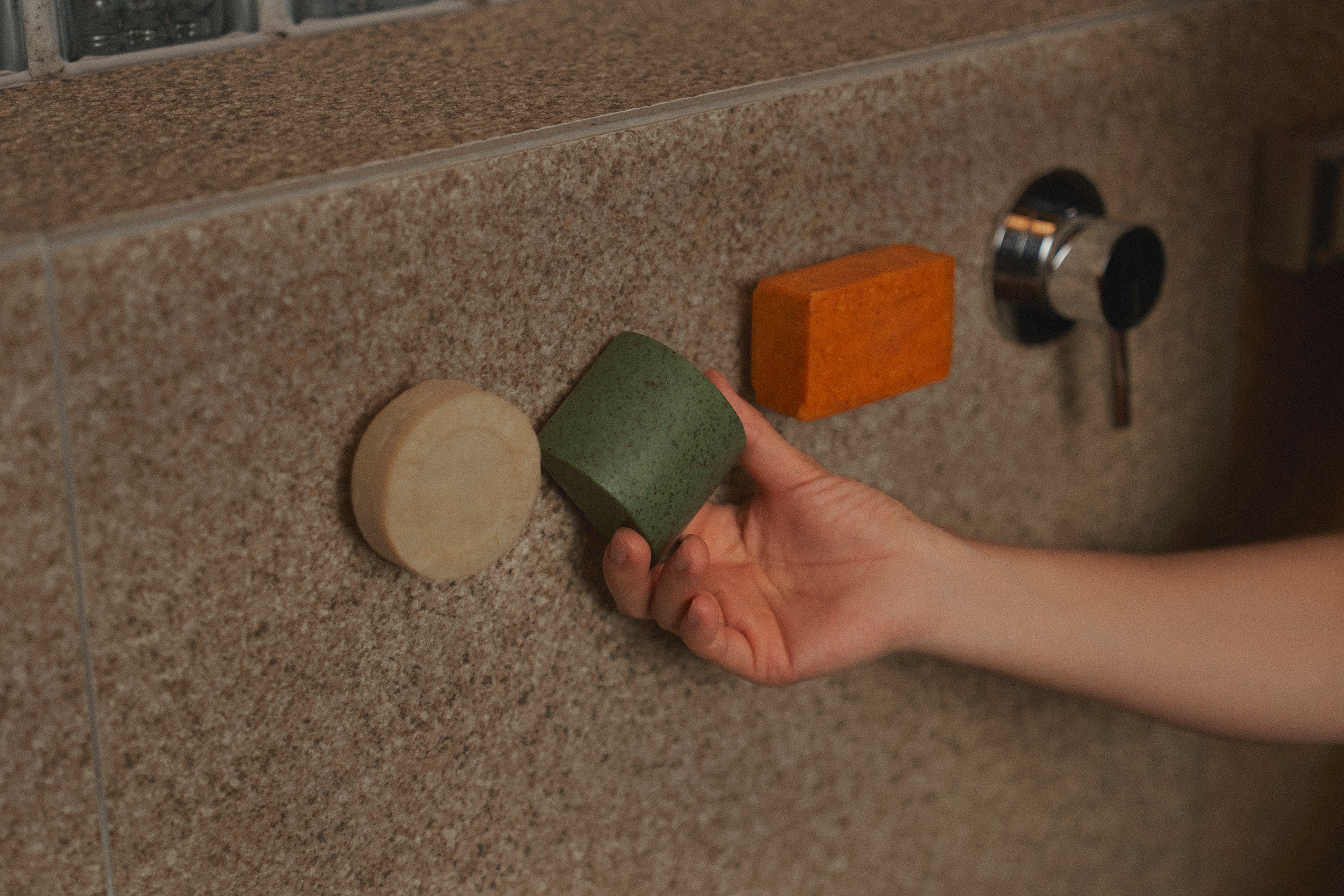 Nhuka Stem magnetic bar holder with shampoo bar, soap bar, and One Bar suspended neatly in the shower. A hand reaches for the bar, evoking restfulness and simplicity. Highlights our Shower Essentials collection.