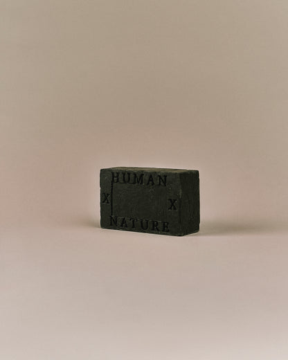 Nhuka Charcoal One Bar shown at an angle with Human x Nature emboss. Handmade in Australia, pH-balanced, sulfate-free, silicone-free, and organic. Warming chai scent with cinnamon, ginger, and nutmeg. Contains pumice for exfoliation. Suitable for hair, face, and body.