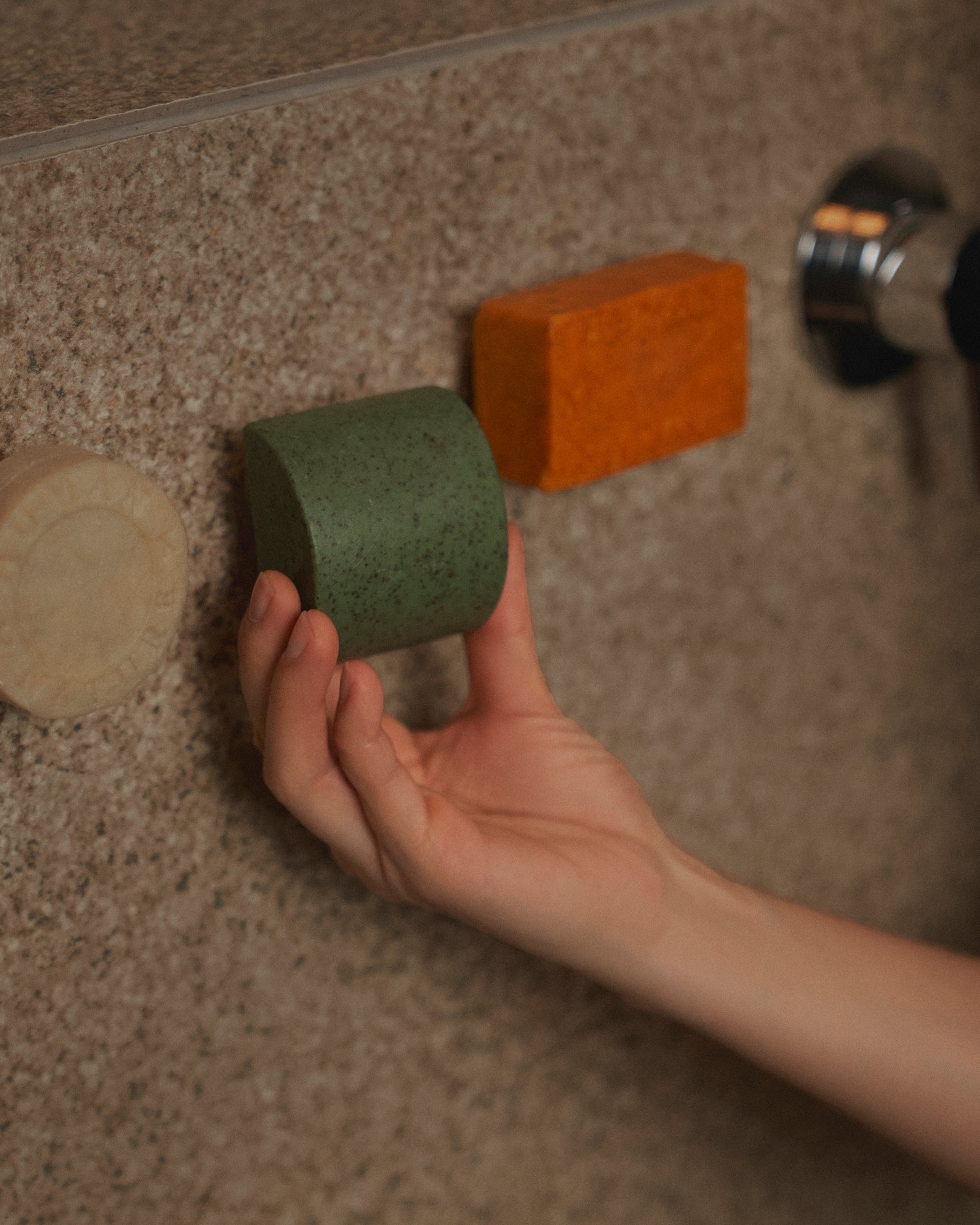 Nhuka Stem magnetic bar holder with shampoo bar, soap bar, and One Bar suspended neatly in the shower. A hand reaches for the bar, evoking restfulness and simplicity. Highlights our Shower Essentials collection.