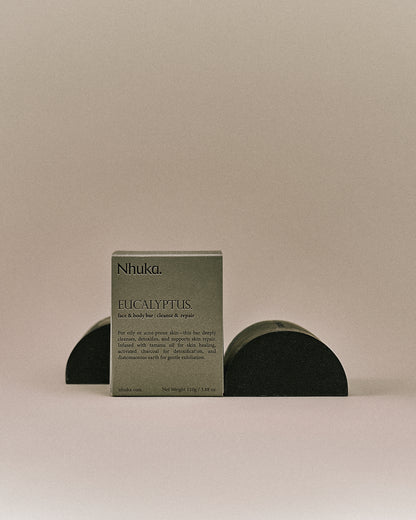 Nhuka organic eucalyptus face and body soap with FSC-certified paper packaging, flanked by two unwrapped bars. Handmade in Australia for cleansing and skin repair. 